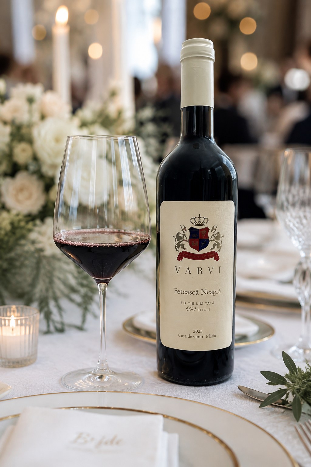 O sticlă de VARVI Fetească Neagră lângă un pahar de vin roșu, pe o masă festivă cu flori albe și lumânări
